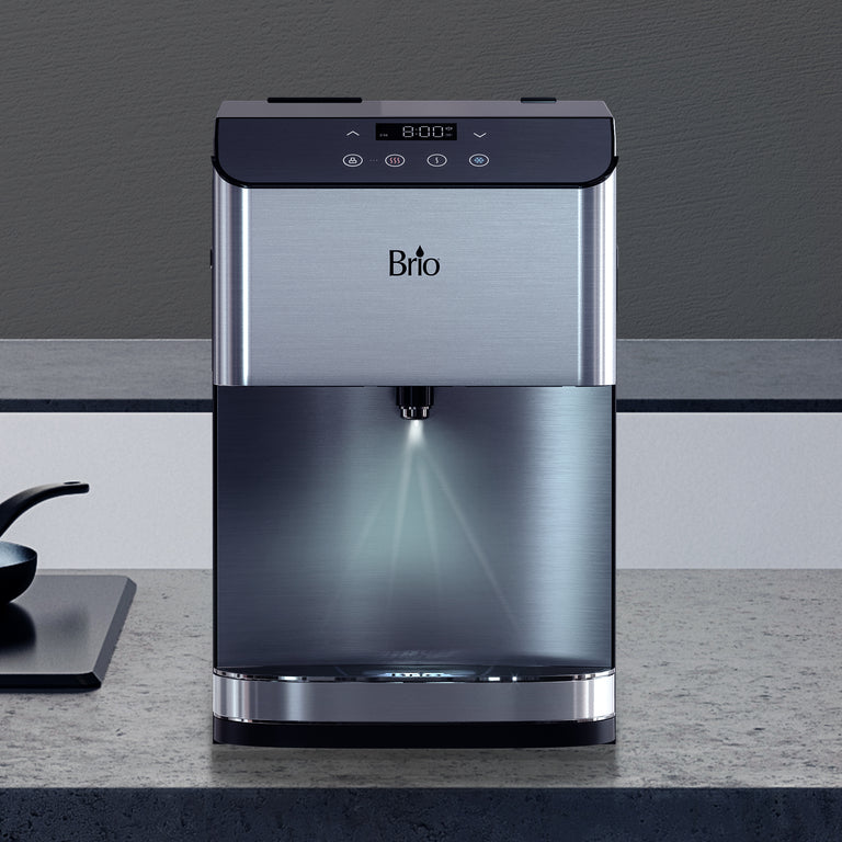 A stainless steel Brio water dispenser sits on a gray countertop. It features a digital display, touch buttons, and a water spout with LED lighting. The background shows a plain gray wall.